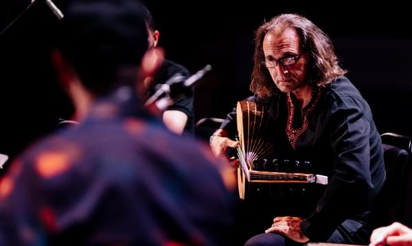 A musician with long hair plays an oud. He looks across at another musician on stage.