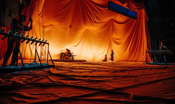 A drummer sits at their kit while someone is sitting on the floor and another is standing, looking at them. The backdrop is a large canvas tent that pillows in dramatic waves above them, lit with a soft warm glow that leaves the rest of the tent in shadow.