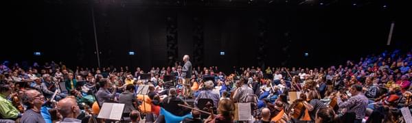 An audience sit on blue and orange beanbags with orchestra players seated on chairs amongst them and a conductor standing in the middle