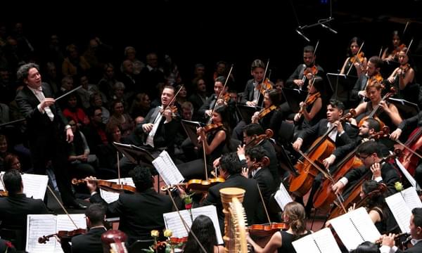 A conductor with curly hair stands on stage conducting an orchestra mid-performance