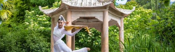 A Chinese dancer holds her instrument and dances in a white costume with flared pants in front of the Chine rotunda at the Edinburgh Botanic Gardens