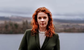 A red-haired woman wearing a dark green jacket stands smiling with a loch and wooded shoreline visible in the distance behind her.