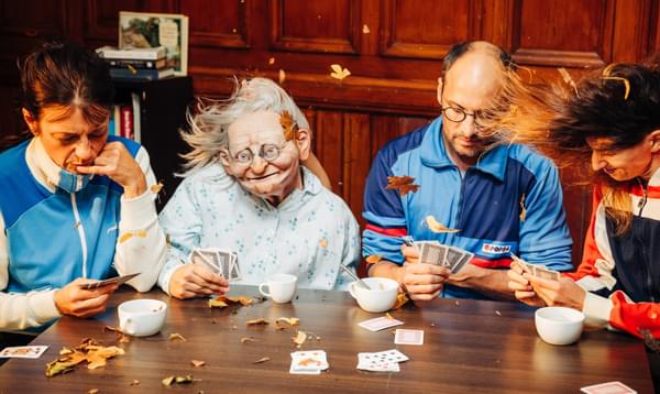 Three actors and a large realistic puppet of an elderly woman play cards and drink coffee as a strong wind blows their hair and orange leaves.