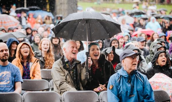 A crowd sing and smile with gentle rain falls, including a couple sharing an umbrella