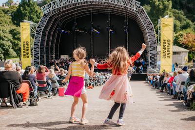 Two young girls earing bright outfits hold hands as they dance in the centre aisle of the Ross Bandstand seating. In the background, a large stage with yellow banners along the sides.