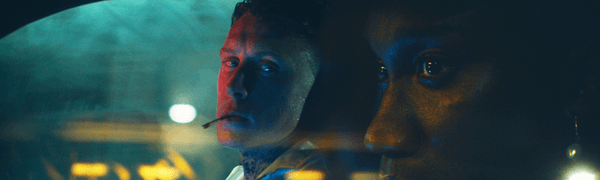 George MacKay (Preston) looks at Nathan Stewart-Jarrett (Jules) in the front seat of a car against dark neon lighting.