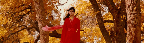 Woman wearing historical red costume holds sword against Autumnal backdrop.
