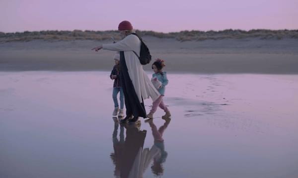 A woman walks with two children and points at something on a pink beach.