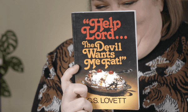 A woman holds a book up to the camera. The title of the book is "Help Lord, The Devil Wants Me Fat!"