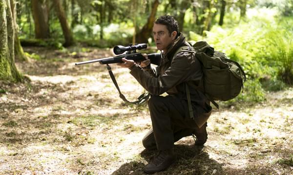 In the woods, a man crouches down and aims a gun