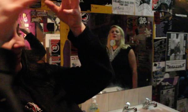 Person looks into mirror in bar bathroom, with posters on surrounding wall.