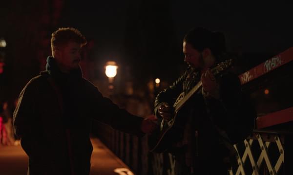 Two men stand in the street at night time, one of them is playing a guitar.