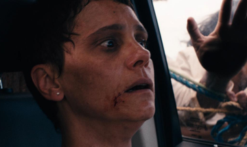 Still from Property. Close-up of a frightened woman sitting in a car with a cut by her mouth. Another person in a baseball cap pushes the vehicle.