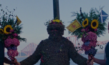 Two men hold flowers and keep the Burryman propped up with the Forth Rail Bridge in the background