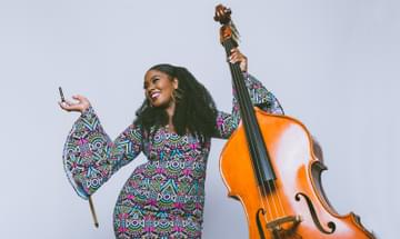 A smiling woman holds a double bass bow casually in her right hand as she looks up and away from the camera, holding up a double bass in her other hand.
