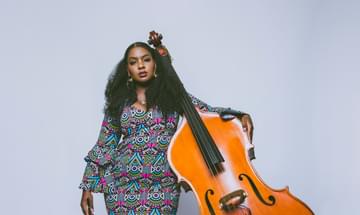 A woman stands with her arm around a double bass, wearing a colourfully patterned mini dress and looking directly at the camera with a steady gaze.