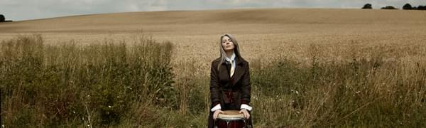 A woman with long grey hair stands in front of a large percussion drum. In the background is a large field and fluffy clouds.
