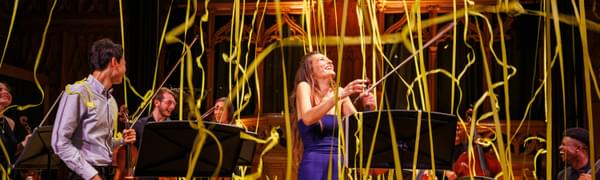Nicola Benedetti, a white woman in a long purple dress, stands holding her violin with her head tipped back and smiling as yellow streamers float down with other musicians surrounding her
