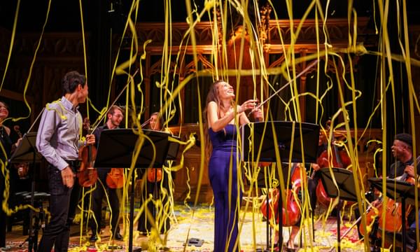 Nicola Benedetti, a white woman in a long purple dress, stands holding her violin with her head tipped back and smiling as yellow streamers float down with other musicians surrounding her