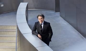 A man with short black curly hair, stands in a suit, leaning against a half wall, outside. The wall is curved, and the building is made from stainless steel.