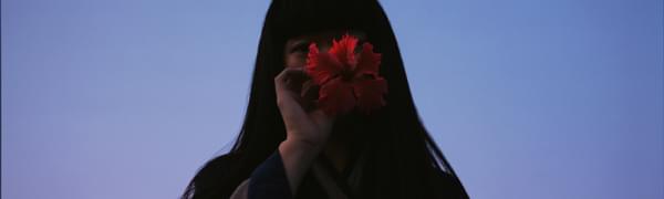 A woman with long black hair covers her face with a hibiscus flower. Behind her, the sky is purple.