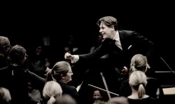Conductor Klaus Makela leans forward and smiles as he conducts the Oslo Philharmonic