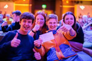 High school students sit on seats in a music venue. They grin holding up a ticket.