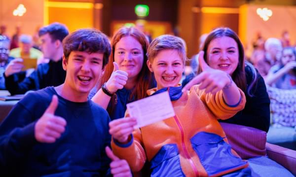 High school students sit on seats in a music venue. They grin holding up a ticket.