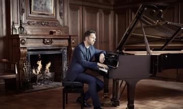 Leif Ove Andsnes sits at a grand piano in an elegant room with wood paneling on the walls and a crackling fire in the fireplace
