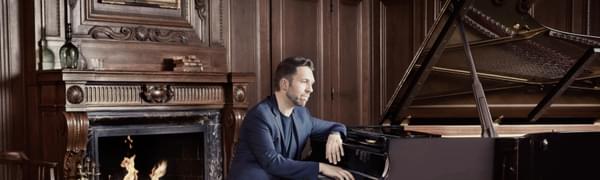 Leif Ove Andsnes sits at a grand piano in an elegant room with wood paneling on the walls and a crackling fire in the fireplace