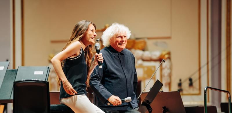 A white woman with long brown hair sits on a stool holding a microphone next to an older white man with white curly hair, both smiling towards an audience.