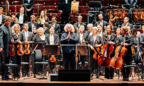 A conductor with big white hair stands to the audience, in front of a large orchestra. He smiles in gratitude