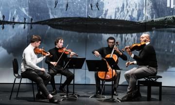 Four young men play instruments as part of a string quartet. They sit on a stage and behind them there is an image of four walkers hiking between lakes and mountains
