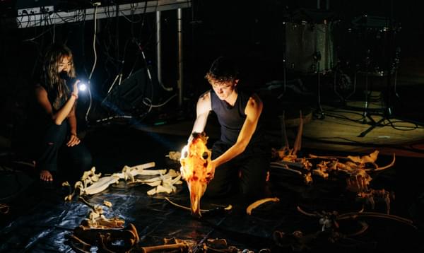 A man in a black tank top holds a horse head skeleton. He is surrounded by horse bones and musical instruments. A woman shines a torch on him.