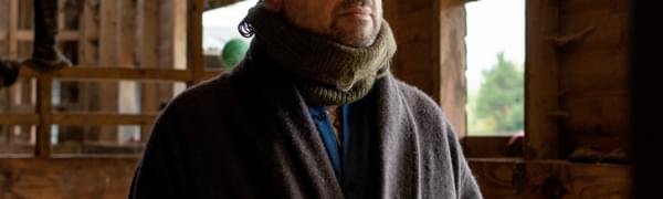A man in a scarf in glasses stands in wooden stables.