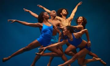 Five dancers jump together in front of a dark blue background. Their movements are graceful and strong.