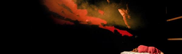 A woman in a red dress sleeps curled up on a bed with her back to the camera, while above her a man descends upside down from a black rock climbing wall, with mystical red smoke billowing around him.