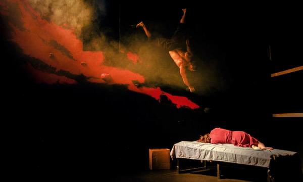 A woman in a red dress sleeps curled up on a bed with her back to the camera, while above her a man descends upside down from a black rock climbing wall, with mystical red smoke billowing around him.