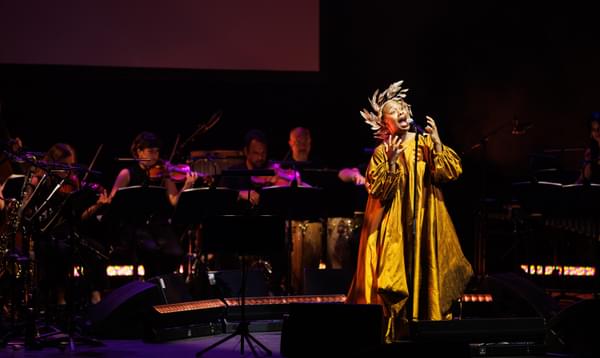 A woman sings in front of a microphone wearing a large gold dress and a gold head piece. Behind her is a band playing instruments.