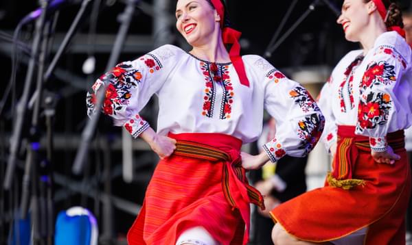 Two women dance and smile on stage, their knees raised and hands on hips. They are wearing traditional dress, with embroidered blouses and red skirts.