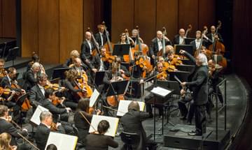 The strings section of the Deutsche Oper Berlin are being conducted by a man with white long hair to the left of the image.