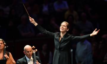 A conductor stands with the conductors baton, arms outstretched and her eyes closed. Next to her sit two strings players.