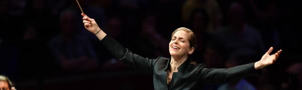 A conductor stands with the conductors baton, arms outstretched and her eyes closed. Next to her sit two strings players.
