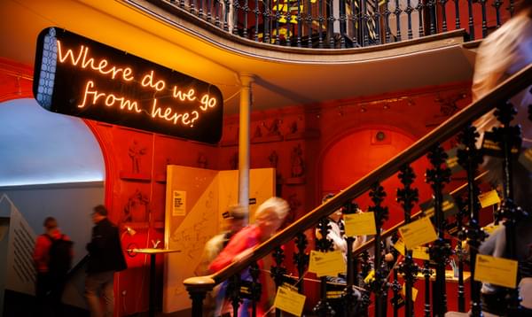 A yellow neon sign says 'Where do we go from here?' as people walk past and up stairs with decorative railings. On the railings are yellow postcards hanging from twine.