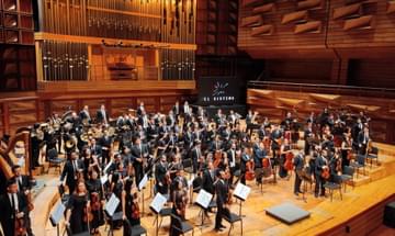 The whole orchestra stands up on an imposing wooden stage. They are all dressed in black, smart clothes and hold their instruments to their chests.