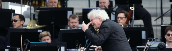 A white-haired man captured vigorously conducting an orchestra, turning to the site to gesture with his left hand. String and wind instrument players are visible behind music stands around him.