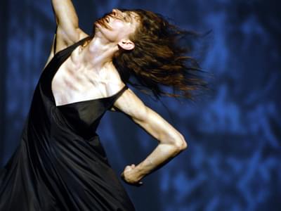 A female dancer in a black dress stretches one arm up, and one arm behind her. She is in motion, her hair flying behind her. Her face is pained, her mouth slightly open.