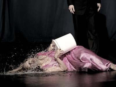 A woman in a pink silk dress lies on the ground and pours a white bucket of water over herself. Behind her, a person in all black stands motionless.