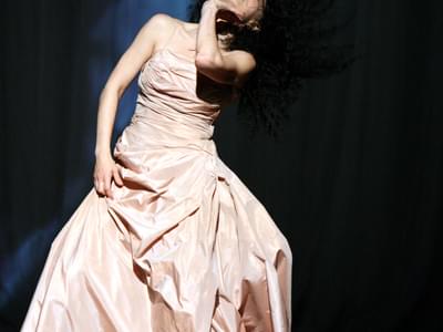 A female dancer in a light pink dress flips her head back, her hair is in motion. Her hand is holding her throat and her expression is pained.