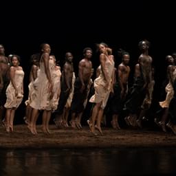 A group of dancers jump with their arms held straight against their legs in white dresses and black trousers above a floor covered in peat